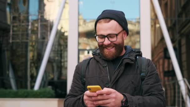 Uomo barbuto in occhiali digitando messaggio e ridendo mentre guardava lo schermo e da parte. Bel ragazzo che usa lo smartphone per chattare sui social media mentre cammina per strada. — Video Stock
