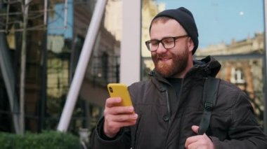 30 'lu yaşlardaki yakışıklı sakallı adam telefon ekranına bakıyor ve sokakta yürürken gülüyor. Gözlüklü neşeli adam komik mesajlar okurken akıllı telefon kullanıyor. Duyguların kavramı.