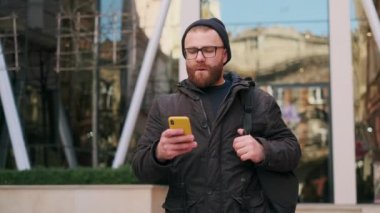 Gözlüklü sakallı adam şok edici bir ifade takınıyor ve akıllı telefon kullanırken hayal kırıklığına uğramış görünüyor. 30 'lu yaşlarda şehir sokağında dikilirken kötü haberler okuyan ciddi bir adam. Duyguların kavramı.