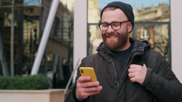 Widok na brodatego człowieka w okularach patrząc na ekran telefonu i śmiejąc się podczas spaceru po ulicy. Przystojny facet przewijanie social media kanał wiadomości podczas korzystania ze smartfona. — Wideo stockowe