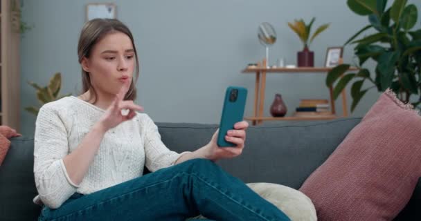 Κωφή γυναίκα χιλιετηρίδας που έχει βιντεοκλήση και δείχνει με νοηματική φράση Τρομερά νέα, ενώ κάθεται στον καναπέ. Γυναίκα πρόσωπο που αναζητούν λυπημένος κατά τη χρήση smartphone για την επικοινωνία. — Αρχείο Βίντεο