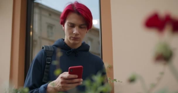 Vista de cerca del hombre hipster con la mochila en su hombro desplazándose y mirando la pantalla del teléfono. Millennial chico con el pelo rojo teñido usando el móvil mientras está de pie en la calle. — Vídeo de stock