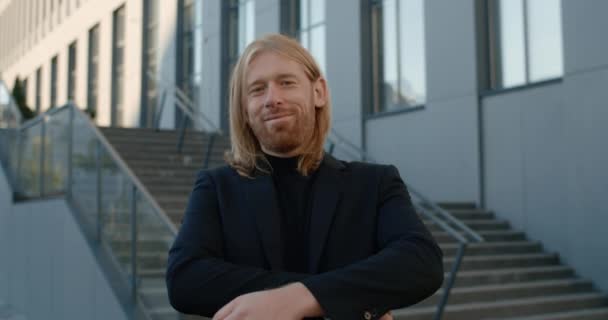 Portrait of happy businessman with long hair crossing arms and looking to camera. Crop view of confident bearded man in suit smiling while standing at stairs outdoors. Concept of success. — Stock Video