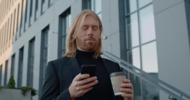 Vista inferior de hombre de negocios guapo beber café de la taza de papel y el uso de teléfono inteligente. Barbudo hombre con el pelo largo comprobar el contenido del teléfono y la pantalla de desplazamiento whle de pie en la calle. — Vídeos de Stock