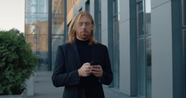 Vista de la cosecha de buen hombre de negocios utilizando el teléfono inteligente y mirando a un lado. Hombre positivo exitoso tocando y presionando la pantalla del teléfono mientras camina en la calle en el área de negocios. — Vídeos de Stock