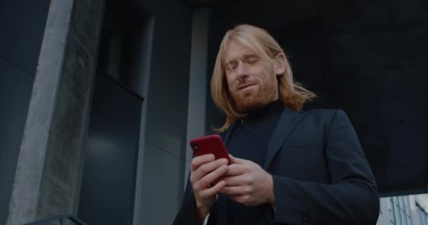Portret przystojnego brodatego mężczyzny używającego smartfona i uśmiechającego się. Widok dolny biznesmena przewijającego i dotykającego ekranu stojącego na zewnątrz. Koncepcja ludzi sukcesu. — Wideo stockowe