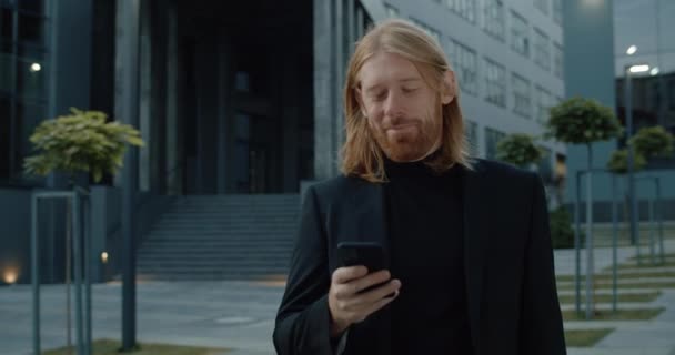 Crop view of long haired man in 30s smiling and looking at smartphone screen while walking. Crop view of handsome happy businessman using phone at street. Concept of successful people. — Stock Video