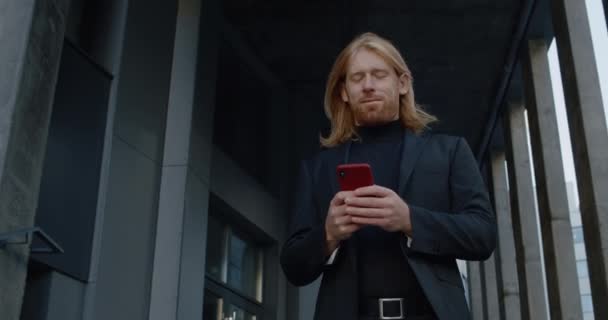 Retrato de un hombre barbudo bien parecido usando un teléfono inteligente y sonriendo. Vista inferior del hombre de negocios escribiendo y tocando la pantalla mientras está parado al aire libre. Concepto de personas exitosas. — Vídeo de stock