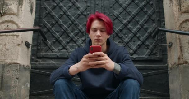 Vue de face de l'homme hipster en vêtements décontractés naviguant sur Internet et regardant l'écran du téléphone. Millennial gars avec les cheveux rouges teints en utilisant smartphone et assis à l'escalier du vieil immeuble. — Video