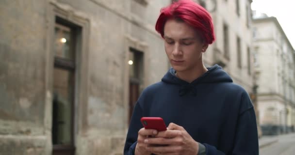 Widok na młodego człowieka z kolczykiem wpisując wiadomość i patrząc na ekranie telefonu. Millennial mężczyzna hipster z farbowanymi czerwonymi włosami za pomocą smartfona podczas spaceru na ulicy starego miasta. — Wideo stockowe