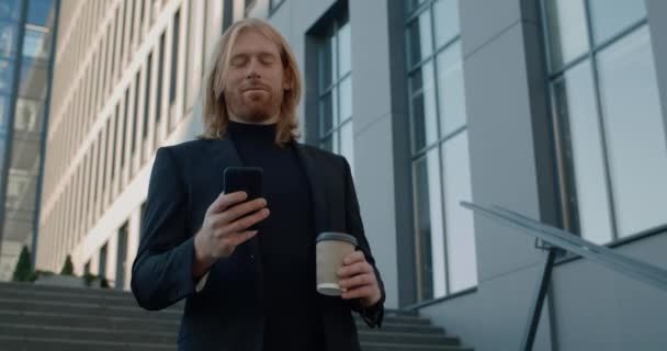 Vista inferior del hombre barbudo con taza de papel de café sonriendo mientras usa el teléfono móvil. Ejecutivo de buen aspecto en traje de navegar por Internet y mirando hacia el futuro mientras baja escaleras. — Vídeos de Stock