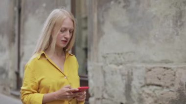 Güzel sarışın kadın eski şehir caddesinde yürürken telefon ekranına bakıyor ve dokunuyor. Akıllı telefon kullanırken internette gezinen zeki bir kadın. İletişim kavramı.