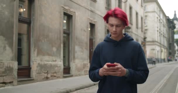 Beskär syn på ung kille med färgat rött hår skriva och titta på telefonskärmen. Millennial manliga hipster i casual kläder med hjälp av smartphone för att chatta på sociala medier när du går. — Stockvideo