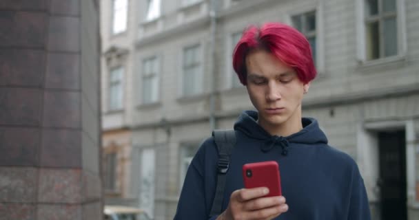 Zamknij widok tysiąclecia facet za pomocą smartfona podczas stania na ulicy. Hipster mężczyzna osoba z farbowanymi czerwonymi włosami wpisując wiadomość i patrząc na ekranie podczas komunikacji w mediach społecznościowych. — Wideo stockowe