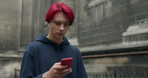 Fiatal férfi hipszter vezeték nélküli fülhallgatóban üzenetet ír miközben nézi és megérinti a telefon képernyőjét. Millenniumi srác tetoválásokkal és festett vörös haj okostelefon használata közben séta az utcán.