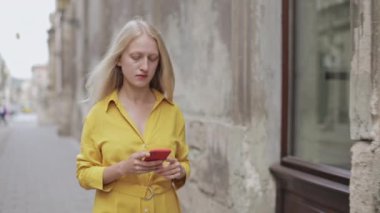 Eski şehir caddesinde yürürken telefon ekranına bakan doğal sarışın kadın manzarası. Genç bayan ciddi kişi akıllı telefon kullanırken internette geziniyor.