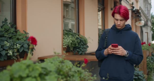 Chlápek v tmavé mikině s taškou na rameni a mobilním telefonem, když stojí na ulici. Muž tisíciletý hipster s barvenými rudými vlasy procházení internetu při psaní a pohledu na obrazovce telefonu. — Stock video