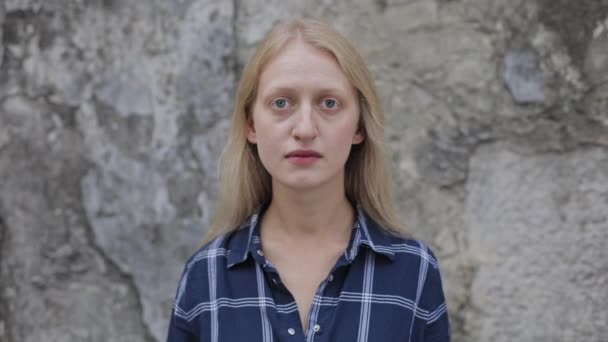 Crop view van jonge blonde haar vrouw op zoek naar camera. Portret van een vrouw in een blauw hemd die poseert terwijl ze buiten staat. Oude stenen muur op de achtergrond. Concept van het echte leven. — Stockvideo