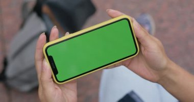 Sokakta otururken video izlemek için telefon kullanan bir kadın görüntüsü. Akıllı telefonu elinde tutan kadın şablon ekranı yatay olarak yapıyor. Krom anahtar kavramı, yeşil ekran.