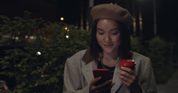 Портрет хорошої молодої жінки, яка використовує смартфон, стоячи на нічній вулиці. Міленіалка в Береті прокручує соціальні медіа стрічка новин і посміхається, тримаючи паперову чашку кави . — стокове відео