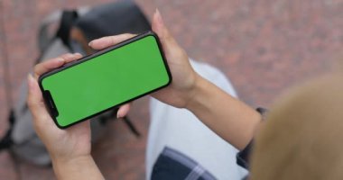 Sokakta otururken video izlemek için telefon kullanan bir kadının en iyi görüntüsü. Yeşil şablon ekranlı akıllı telefonu yatay olarak tutan kadın. Krom anahtar kavramı, modelle.