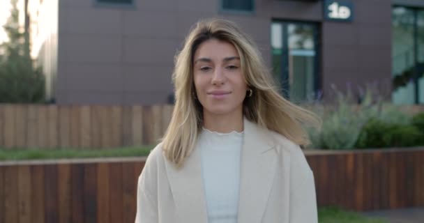 Retrato de mujer joven atractiva con el pelo rubio mirando y posando a la cámara. Vista de la cosecha de la persona femenina alegre milenaria sonriendo mientras está de pie en la calle. Acercar. — Vídeo de stock