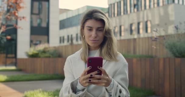 Positiv ung kvinna skriva på smartphone skärmen när du sitter på bänken. Vacker kvinnlig person som surfar på internet och tittar på kameran med leende på stadens gata.Zooma in. — Stockvideo