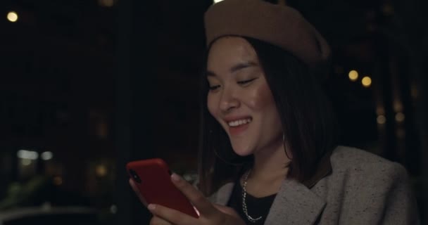 Närbild av glad kvinnlig person kontrollera smartphone innehåll och skriva meddelande när du går på natten gatan. Ung kvinna i snygg basker ler när du använder telefon. — Stockvideo
