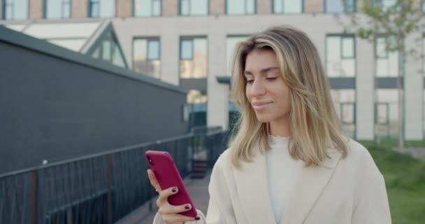 Mulher milenar atraente com cabelo loiro usando smartphone enquanto caminha na rua da cidade. Visualização da colheita da pessoa feminina feliz sorrindo enquanto rola o feed de notícias das mídias sociais. — Vídeo de Stock