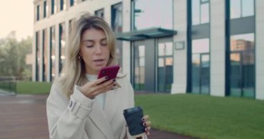 Sarı saçlı, akıllı telefon ses tanıma cihazı kullanan, kağıt kahve fincanı taşıyan genç bir kadının yakın görüntüsü. Başarılı bir kadın sokakta yürürken konuşuyor ve uygulama kullanıyor..