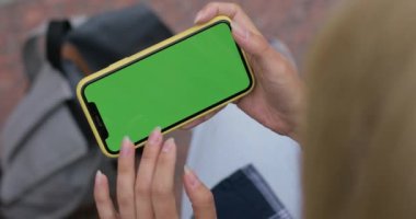 Sarışın kadının açık havada otururken video izleyip şablon görüntüsü. Yeşil ekrana dokunurken ve kaydırırken akıllı telefonu yatay olarak tutan kadın.