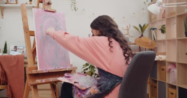 Bruna dai capelli lunghi ragazza pittura quadro moderno mentre seduto di fronte a molbert. Adolescente talentuoso creazione durante l'applicazione di vernice e facendo colpi su canva. A casa. Concetto di arte. — Video Stock