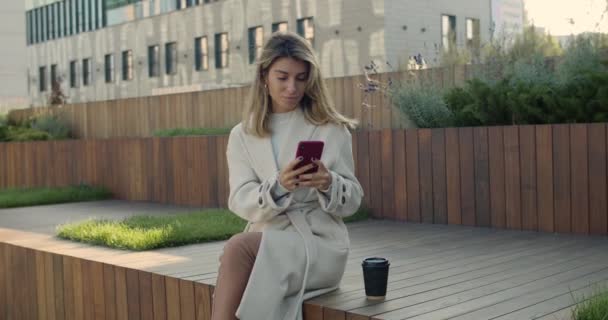 Stylish woman typing at smartphone screen while sitting on bench near paper coffee cup. Attractive female person browsing internet while spending free time at city street. — Stock Video