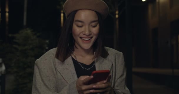 Vista de corte da pessoa feminina atraente usando smartphone enquanto caminha na rua noturna. Jovem mulher alegre na boina rindo e olhando para a tela enquanto tem mensagem engraçada. — Vídeo de Stock