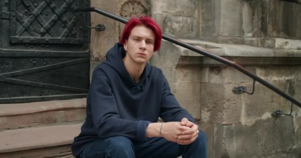 Portret tysiącletniego hipstera z farbowanymi rudymi włosami patrzącego w kamerę. Widok na młodego człowieka z kolczykiem i tatuażami w ubraniu casual siedzi na schodach starego budynku. — Wideo stockowe