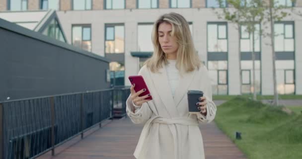 Crop view van gelukkige vrouw met blond haar met behulp van smartphone en zeggen wow whle dragen papieren koffiebeker. Succesvolle vrouwelijke persoon met goed nieuws tijdens het lopen op straat. — Stockvideo