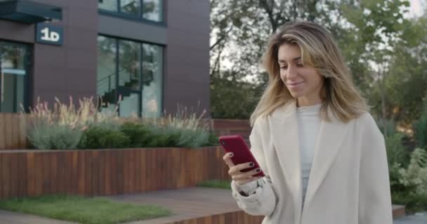 O femeie tânără atractivă care folosește smartphone-ul în timp ce zâmbește și merge pe stradă. O femeie elegantă defilează ecranul telefonului și mângâie părul în timp ce vântul îi suflă pe față . — Videoclip de stoc