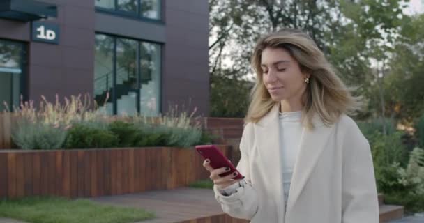 Mujer de negocios alegre usando teléfono inteligente mientras camina por la calle de la ciudad. Vista de la cosecha de la mujer elegante sonriendo y desplazándose por la pantalla del teléfono durante la navegación interent. Concepto de comunicación. — Vídeos de Stock