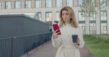 Sarı saçlı, akıllı telefon kullanan ve kağıt kahve fincanından içen bir kadın. Güzel bir kadın sosyal medyada haber yayını yapıyor ve sokakta yürüyor..