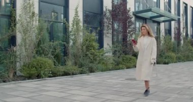 Akıllı telefon kullanan ve gülümseyen gündelik kıyafetli güzel bir genç kadın. Güzel görünümlü, mutlu bir kadın internette gezinirken şehir caddesinde yürürken ve rüzgar saçlarını savururken..