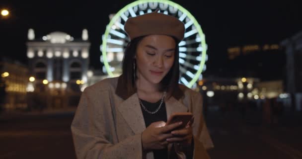 Kvinnlig person som använder smartphone stående på natten gatan. Snygg kvinna höja huvudet och titta på kameran samtidigt skriva meddelande. Pariserhjul med ljus i bakgrunden. Zooma in. — Stockvideo