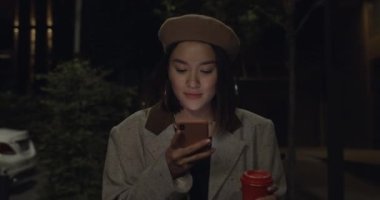Gece sokakta yürürken akıllı telefon kullanan çekici bir genç kadın manzarası. Milenyum tarzı bereli bayan elinde kağıt kahve fincanı tutarken sosyal medya haberleri sunuyor..
