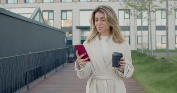 Vista de la cosecha de la mujer de negocios con el pelo rubio con el teléfono inteligente y la celebración de taza de café de papel. Persona femenina exitosa desplazándose por las redes sociales noticias y caminando por la calle. — Vídeo de stock