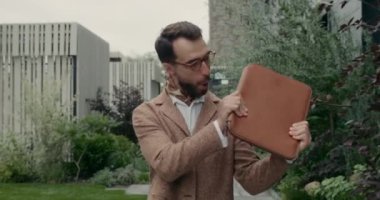 Şehir caddesinde yürürken sallanan ve deri klasörle sarılan gözlüklü heyecanlı adamın görüntüsü. Tarz sahibi sakallı iş adamı gülümsüyor ve mutlu bir başarı elde ediyor..