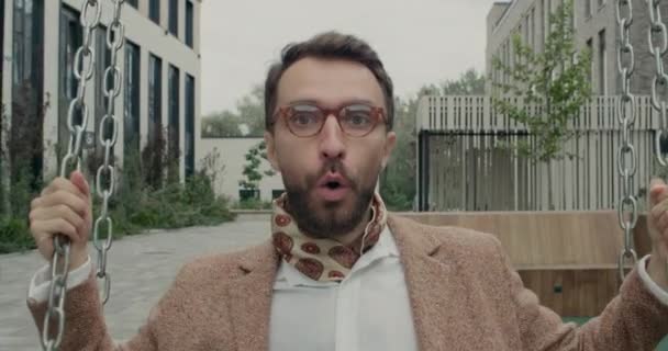 Primer plano del hombre barbudo con gafas balanceándose y riendo mientras mira a la cámara. Feliz hombre de negocios con pañuelo divirtiéndose mientras monta en columpio establecido cerca de edificios de oficinas. — Vídeo de stock