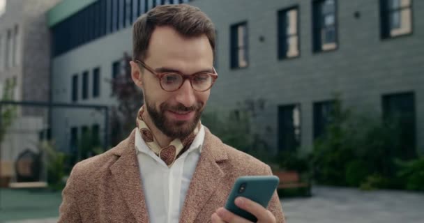 Zblízka pohled veselého podnikatele ve stylovém obleku pomocí smartphonu a s úsměvem. Pohledný muž v brýlích rolování telefonní obrazovku, zatímco stojí na ulici City. Koncept úspěchu. — Stock video