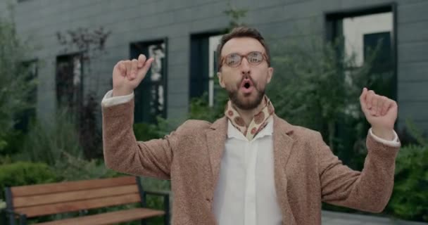 Vzrušený muž v brýlích a krčníku zpívá a cvaká prsty při chůzi po ulici. Stylový vousatý muž hledající kameru a usmívající se při oslavě úspěchu. — Stock video