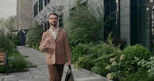 Alegre chico guapo en vasos que llevan bolsa y taza de papel de café. Elegante hombre de negocios barbudo con cuello y traje sonriendo mientras camina por la calle cerca del edificio de oficinas. — Vídeo de stock