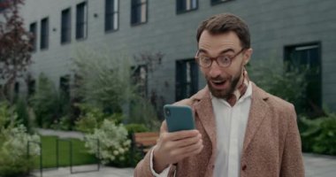 Şık giyinmiş mutlu bir adamın akıllı telefon kullanarak heyecanlı bir yüz ifadesi takınması ve başarıyı kutlaması. Gözlüklü adam telefon ekranını kaydırıyor ve sokakta yürürken seviniyor..