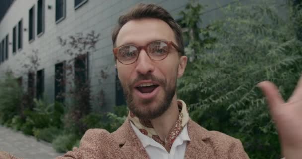 Vista de cerca del hombre de negocios guapo en gafas mirando a la cámara y hablando. Vista de la cámara del teléfono inteligente del hombre barbudo con el pañuelo sonriendo y saludando mientras tiene videollamada. Al aire libre. — Vídeo de stock